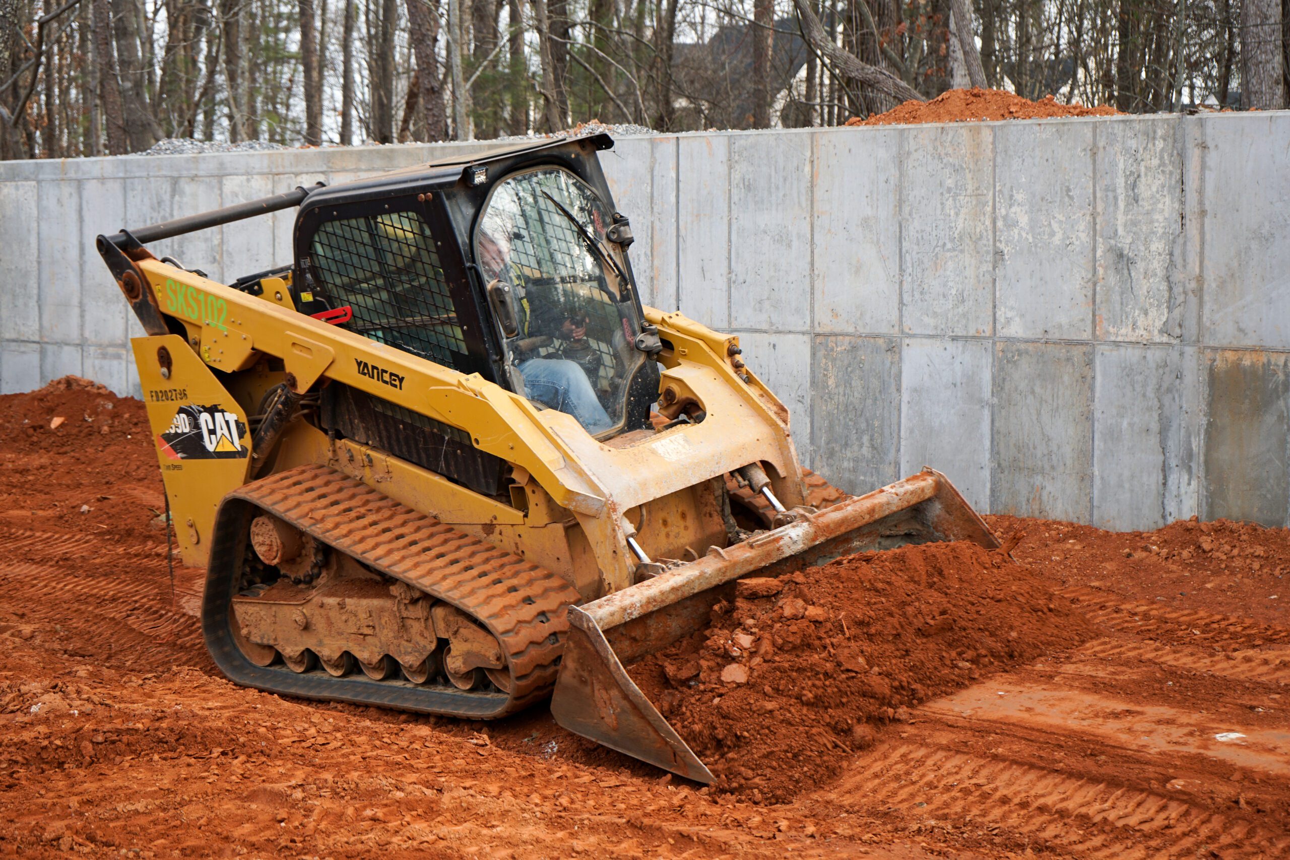 CAT compact track loader grading red clay soil beside a concrete retaining wall at a construction site.